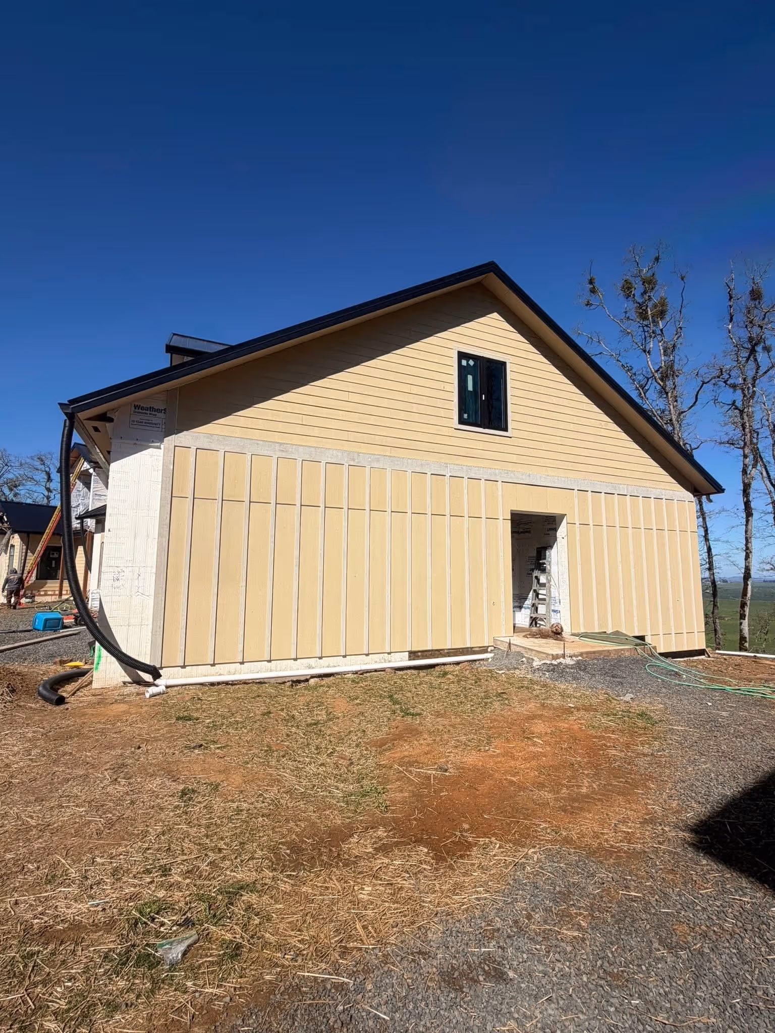 Side view of a partially constructed beige house with vertical siding panels and a single window under a pitched roof against a clear blue sky.