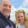 Smiling man and woman posing outdoors with a partly cloudy blue sky background.