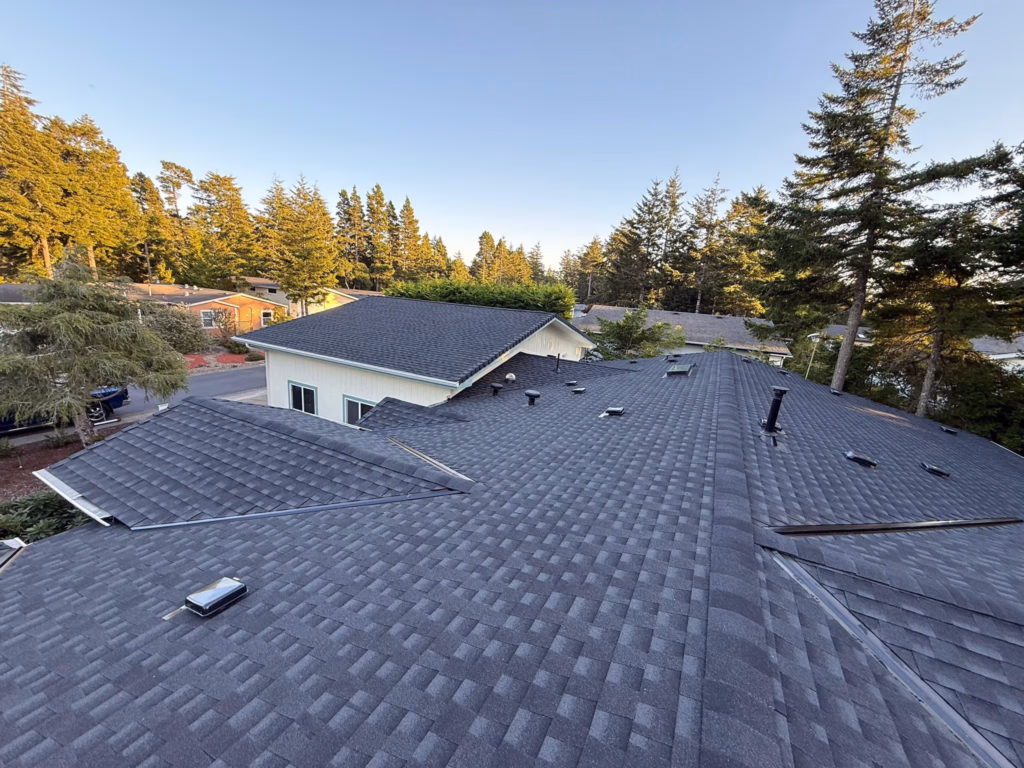 Aerial view of a dark gray shingled residential roof with vent pipes and skylights, surrounded by trees and other houses under a clear sky.